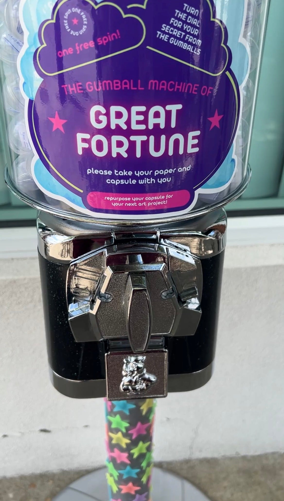 Gumball machine with a purple label reading 'The Gumball Machine of Great Fortune' on a sidewalk. Experiential public art installation by Bailey Lewis, copyright 2026 all rights reserved. 